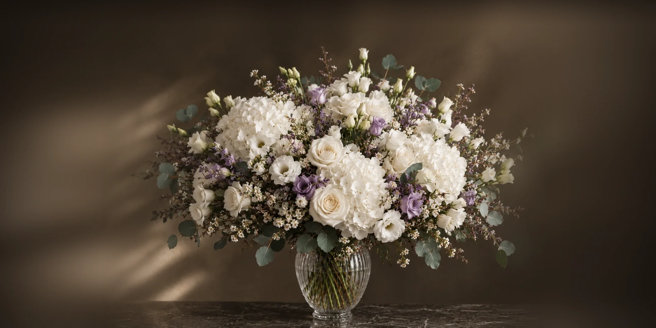 Un grand bouquet de roses blanches, d’hortensias, de lisianthus mauves et d’eucalyptus dans un vase de cristal
